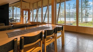 Un comedor amplio con una larga mesa de madera y sillas, ventanas grandes que ofrecen vistas a árboles y un cuerpo de agua.