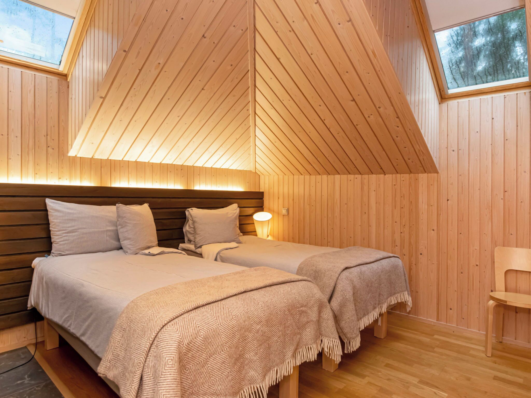 A room with wooden walls and ceiling, two beds with headboards, a bedside table with a lamp, and a window.