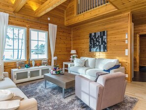 A cozy cabin interior with a wooden ceiling, a white sofa, a pink armchair, a coffee table, and a large window with curtains.