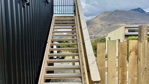 Un escalier en bois mène à un bâtiment à l’extérieur métallique, entouré d’une clôture en bois, avec un paysage montagneux en arrière-plan.