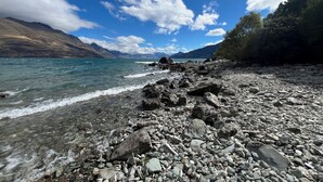 Une côte rocheuse bordée d’une eau claire, avec des montagnes en arrière-plan et un ciel partiellement nuageux.