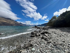 A rocky shoreline with clear water, mountains in the background, and a partly cloudy sky. - Beautiful lake front home (Queenstown)