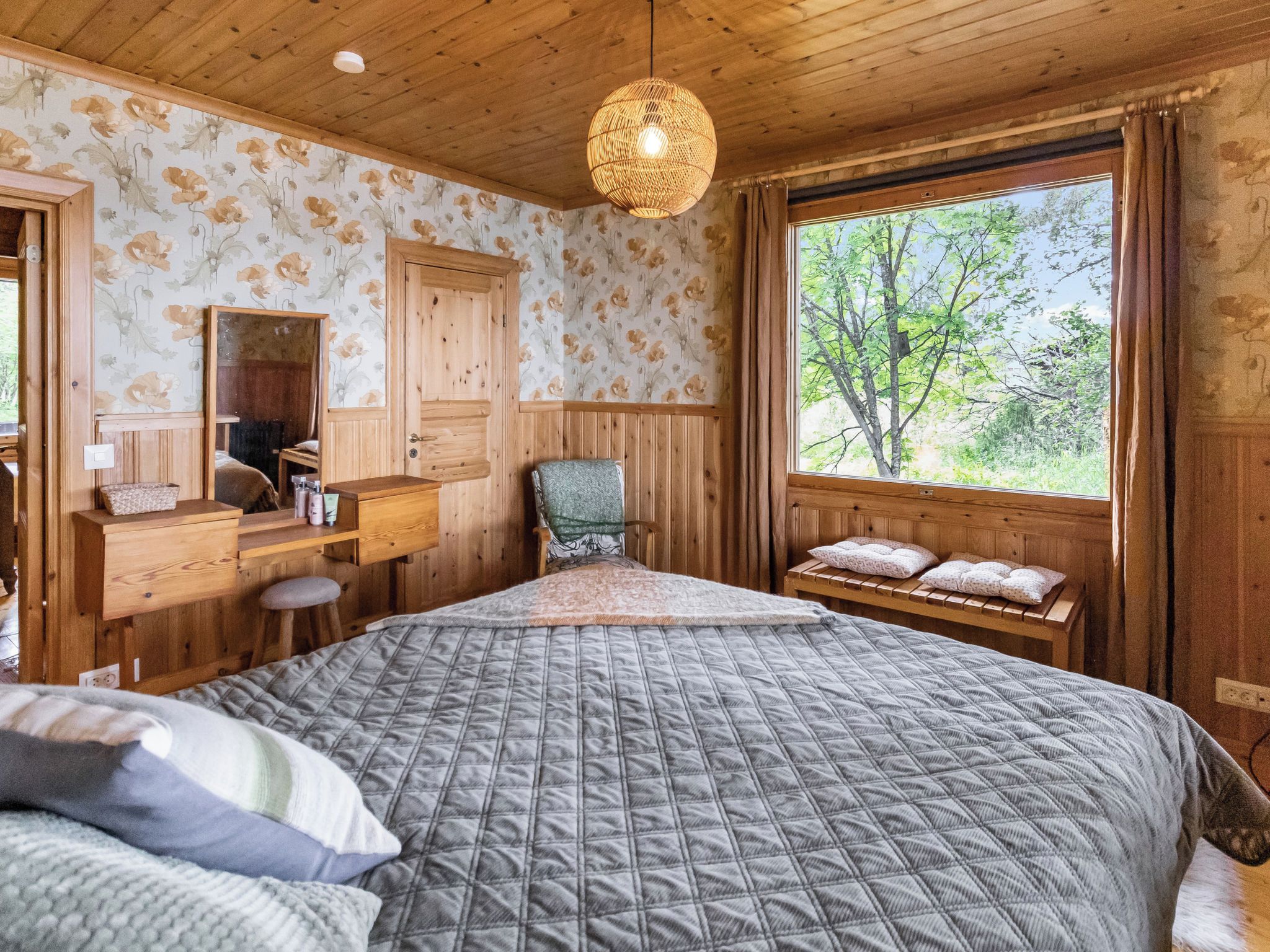 A bedroom with a large bed, a wooden dresser, a chair, and a window with a view of trees.