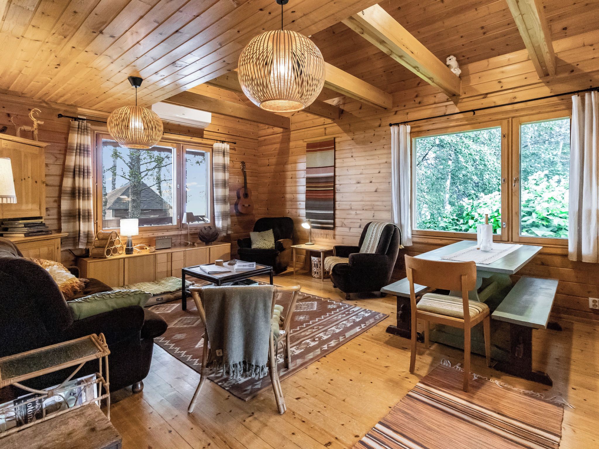 A cozy cabin interior with wooden walls, a dining area, and a large window offering a view of greenery.