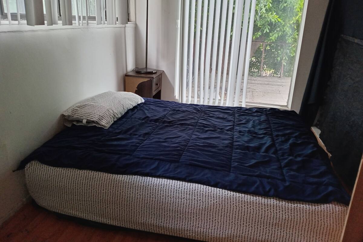 Une chambre avec un lit, une table de chevet, une lampe et une fenêtre avec des stores.