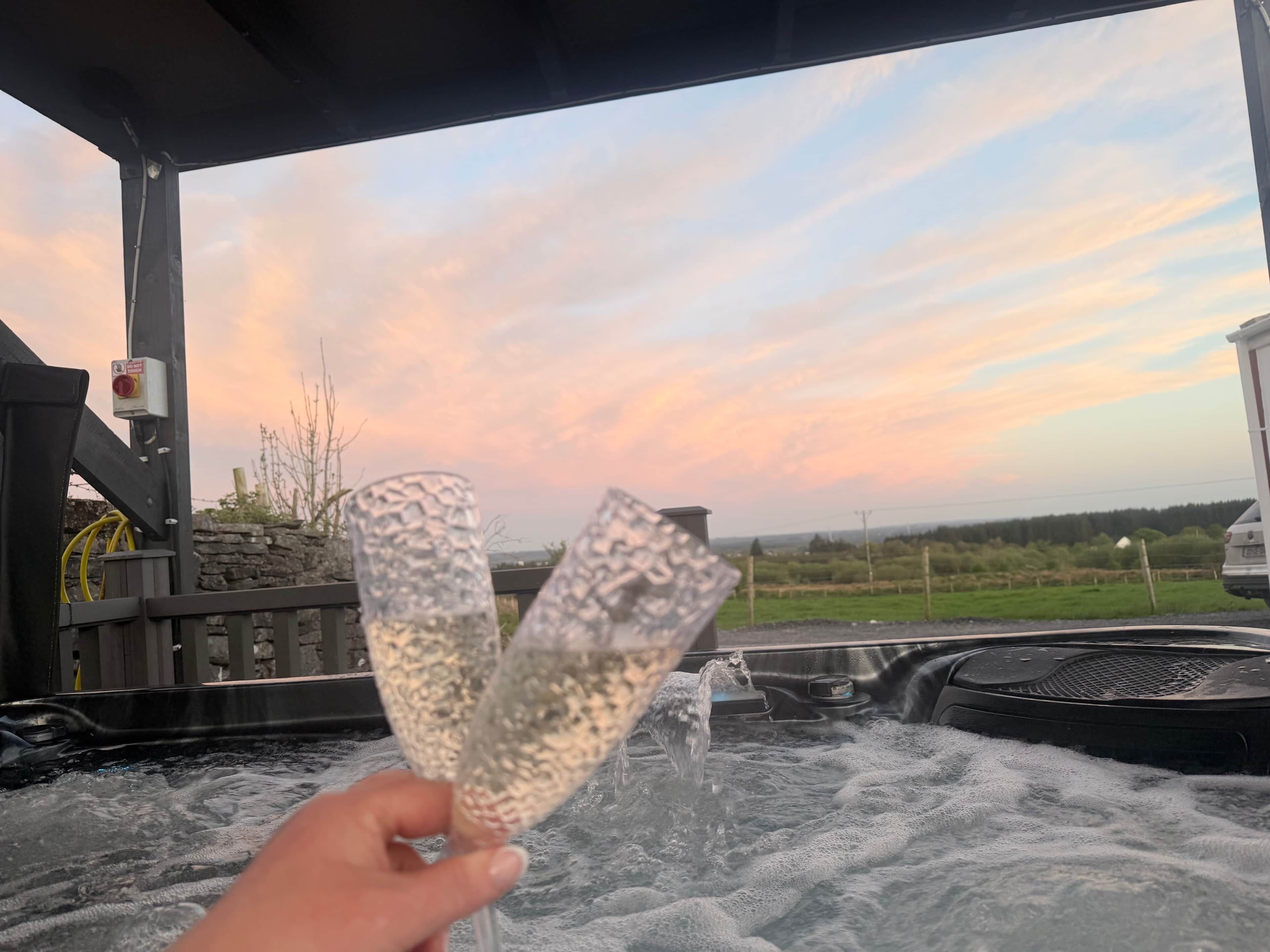 Ett bubbelbad där en person håller i två glas, en stenmur och en vy över solnedgången.