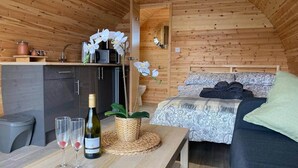 Interior acolhedor de uma cabana com teto de madeira, cama com roupa de cama estampada, mesa de jantar com copos de vinho e uma garrafa, e uma pequena área de cozinha.