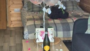 Una habitación acogedora con techo de madera, un sofá, una mesita con una botella y vasos, y un arreglo de globos con un cartel de 'Happy Birthday'.