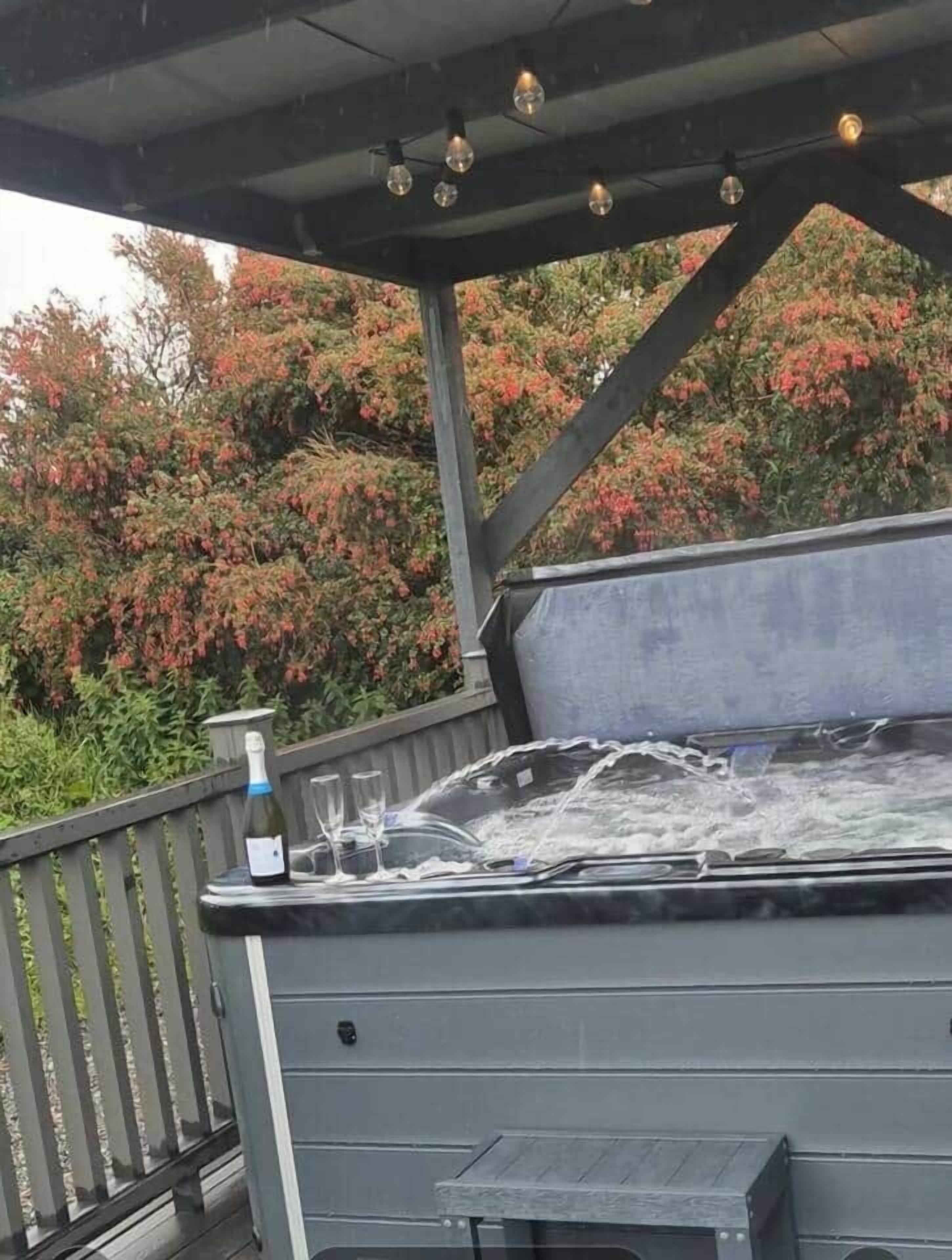 Un bain à remous avec une bouteille et des verres sur une terrasse en bois, entouré du feuillage automnal.