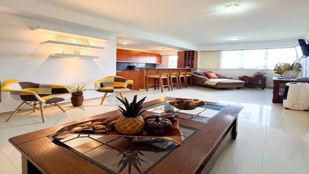 A modern living room with a glass coffee table, a kitchen in the background, and a dining area with chairs.