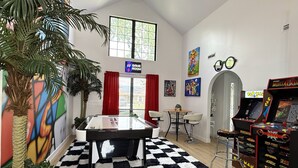 Una habitación con mesa de billar, juegos de arcade, una planta grande y un mural.