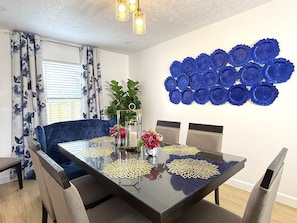 Un coin repas avec une table en verre, des chaises à l’assise bleue, un élément décoratif bleu fixé au mur et une fenêtre ornée de rideaux à motifs floraux.