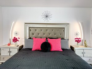 Une chambre à coucher avec une tête de lit capitonnée, une literie à motifs, des coussins décoratifs, des tables de chevet avec des lampes et des œuvres d’art murales.