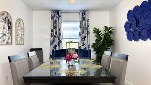 Comedor con mesa de vidrio, sillas tapizadas en gris, sofá azul, planta en maceta y decoración mural.