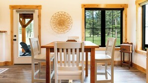 Een eethoek met een houten tafel en stoelen, een decoratief wandobject en grote ramen met uitzicht op groen.