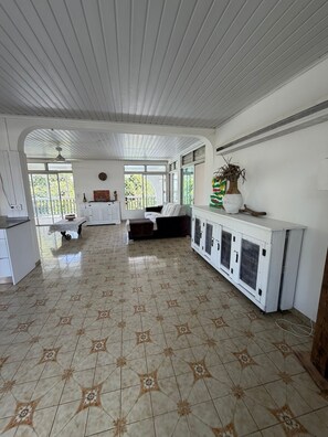 A spacious room with a tiled floor, a sofa, a dining table, and a cabinet with air conditioning units. - The Attic (Le Robert)