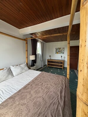 A room with a bed, a bedside table, a lamp, a dresser, and a window with curtains. - The Attic (Le Robert)