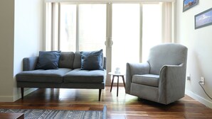 Ein Wohnzimmer mit grauem Sofa und Sessel, Holzboden und großen Fenstern.