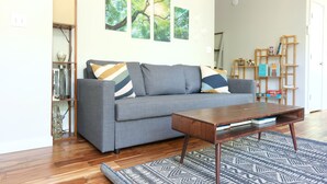 Ein Wohnzimmer mit grauem Sofa, einem Holztisch und einem gemusterten Teppich. An den Wänden hängen drei Bilder von Bäumen.