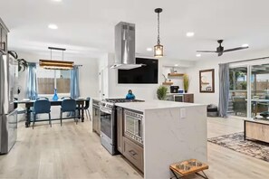 A modern kitchen with a central island, stainless steel appliances, and a dining area with blue chairs. - Designer Escape with Pool, Firepit, Game Room, BBQ (San Diego)