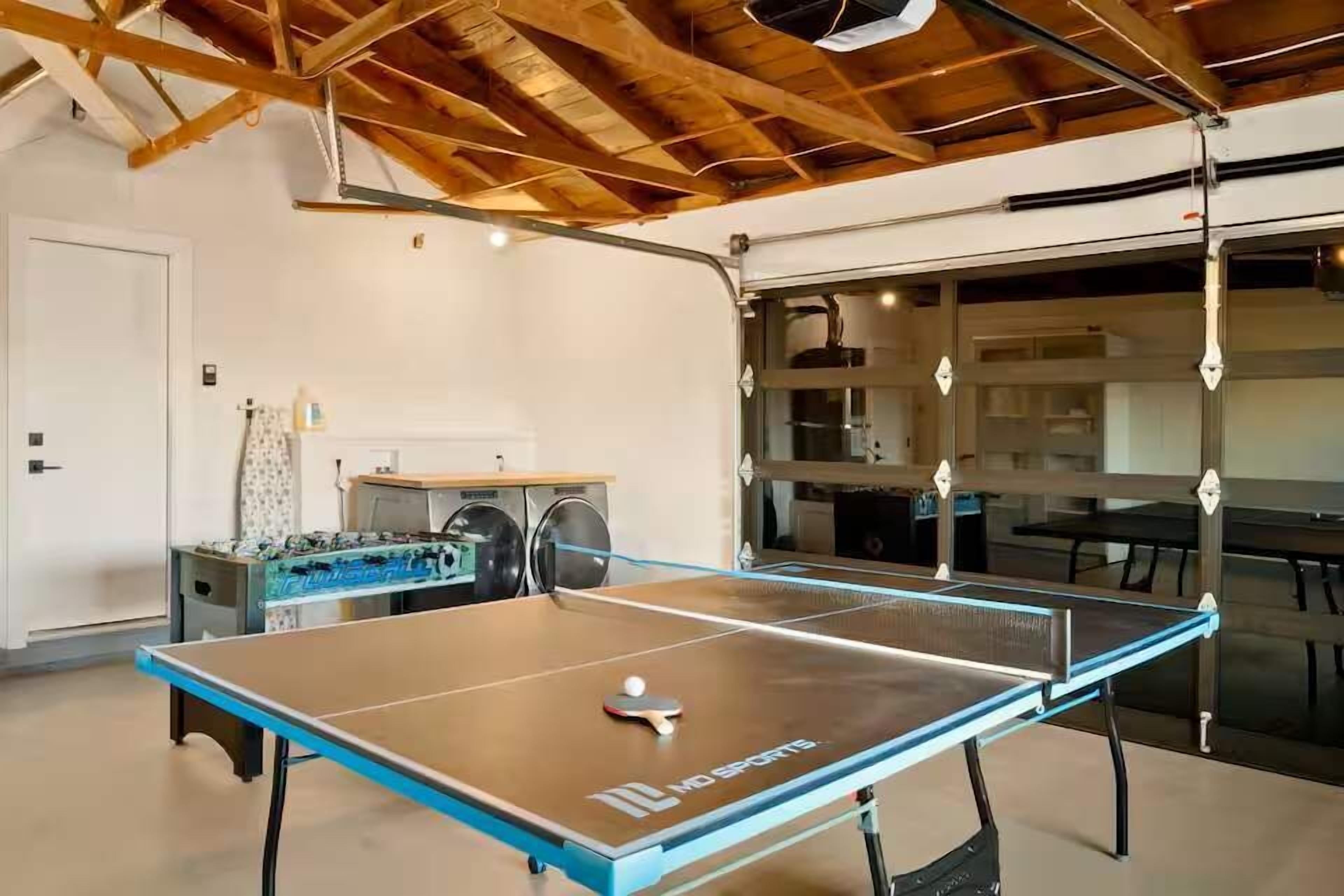 Ein Tischtennistisch mit Ball, ein Kicker und eine Waschgelegenheit in einem Raum mit freiliegenden Holzbalken und einem großen Fenster.