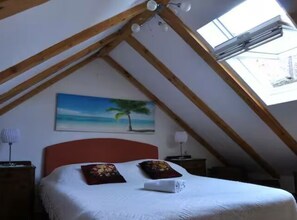 Un dormitorio con techo inclinado, una cama con colcha blanca, dos almohadas, un cuadro de una palmera y un tragaluz.