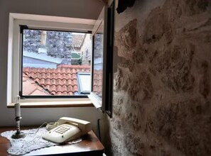 A room with a window overlooking a red-tiled roof, a desk with a telephone, and a wall with a textured surface. - Charming Old Town Getaway (Stari Grad)