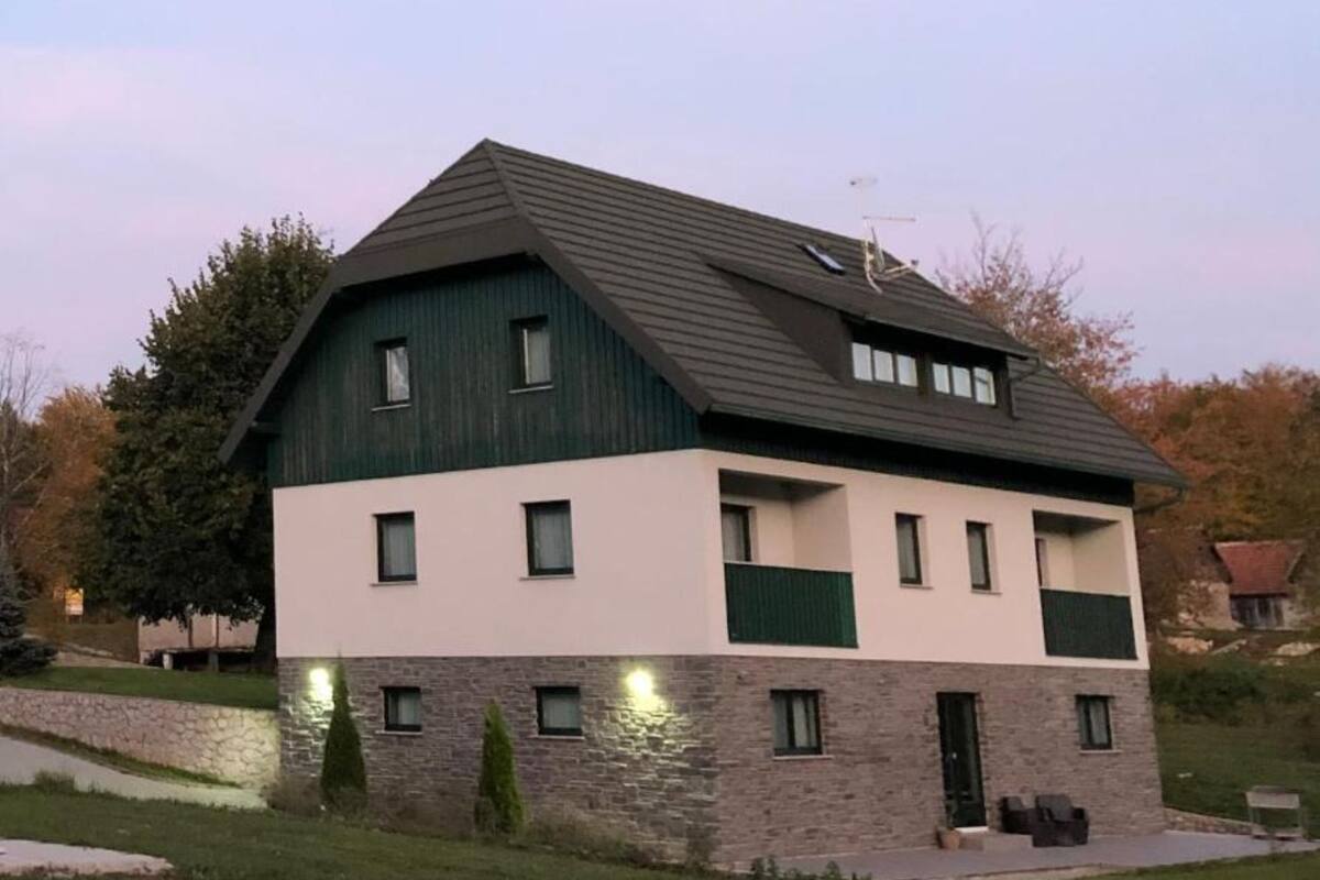 Una casa de dos pisos con base de piedra y techo verde, con varias ventanas y un pequeño porche.