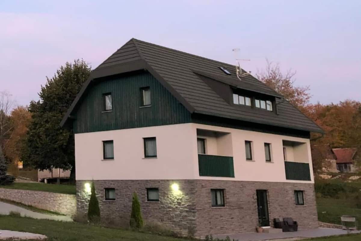 Una casa de dos pisos con base de piedra y techo verde, con varias ventanas y un pequeño porche.