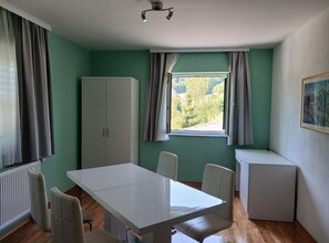 Une pièce avec une table blanche, quatre chaises, un meuble, un radiateur, une fenêtre avec des rideaux et un tableau encadré au mur.