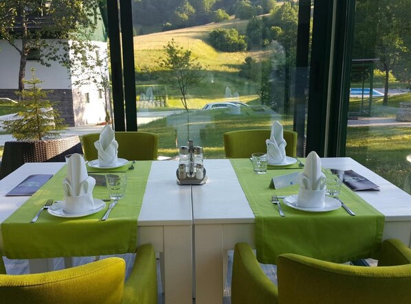 A dining table set for four with green tablecloths, white plates, and yellow chairs, overlooking a scenic landscape. - Villa Lika Annex (Plitvicka Jezera)