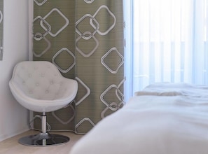 A hotel room with a bed, a white chair, and a patterned curtain.