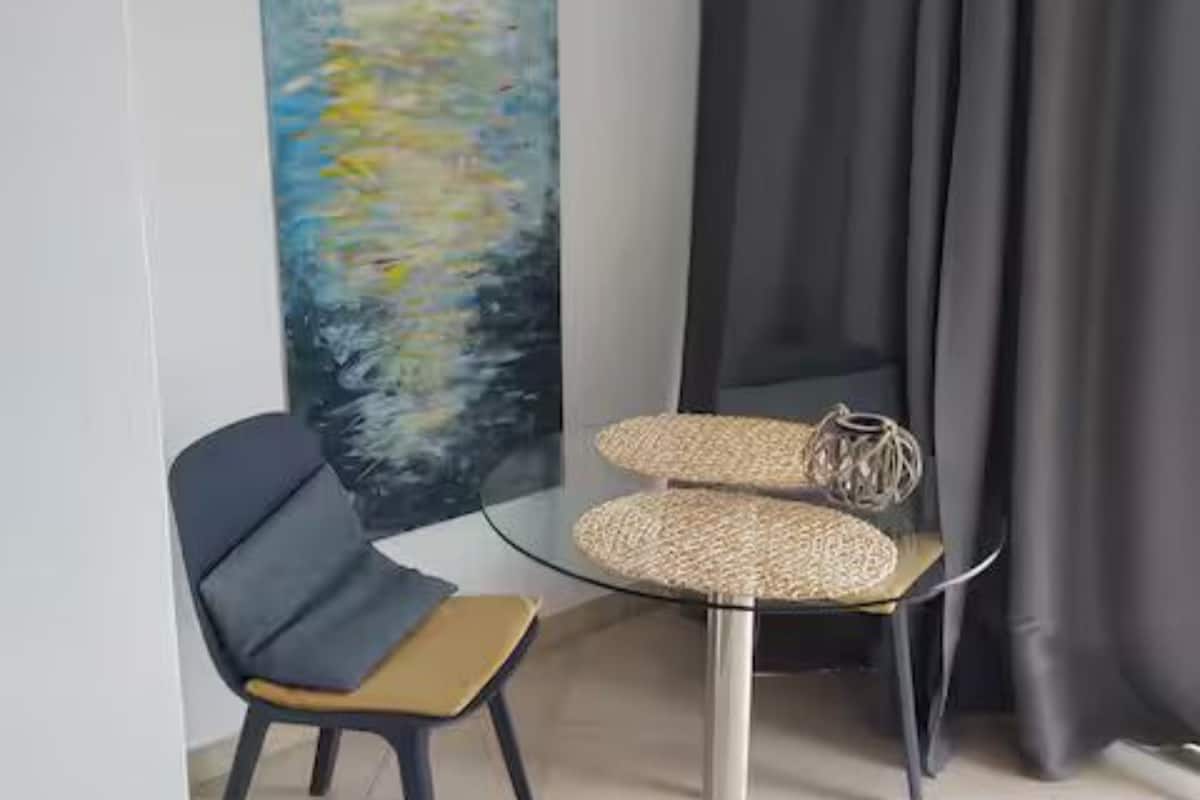 Una habitación moderna con una mesa de vidrio, una silla, un cuadro y una cortina.