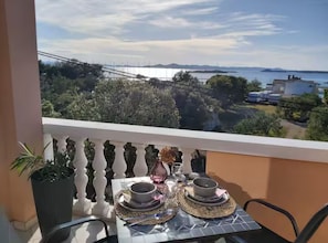 A balcony with a table set for two, overlooking a coastal landscape with trees and buildings.