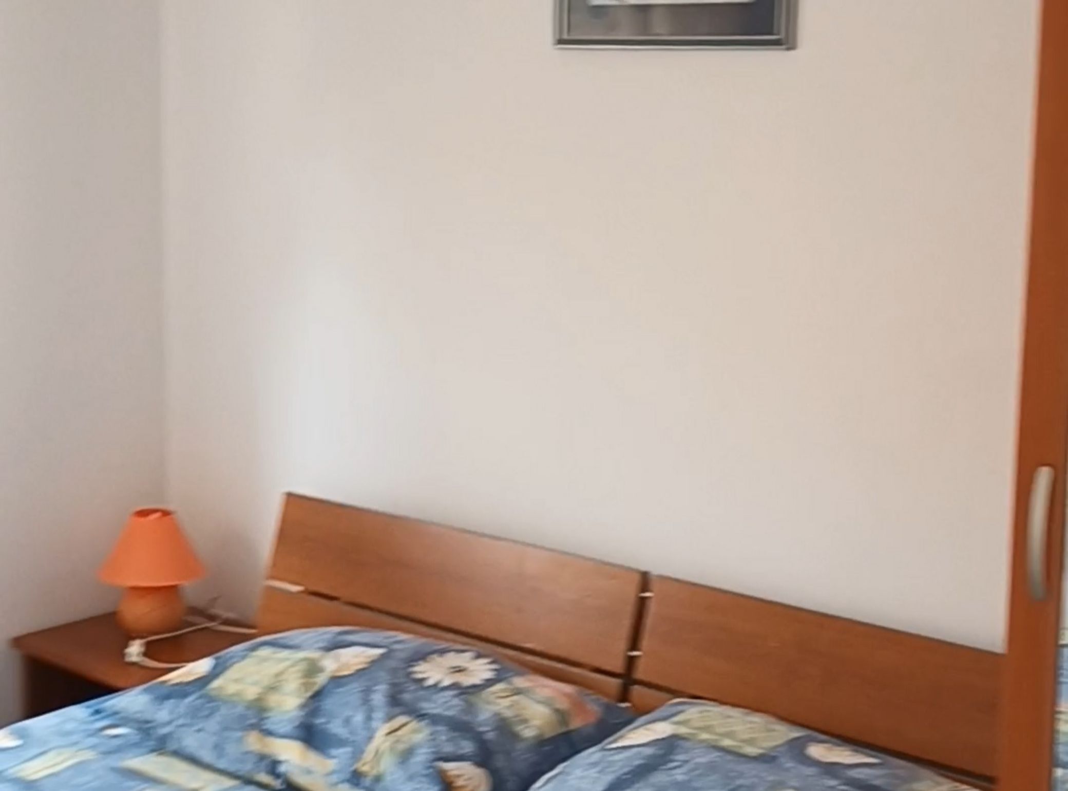 A bedroom with a wooden headboard, a bed with a floral bedspread, a nightstand with an orange lamp, and a wall-mounted light fixture.
