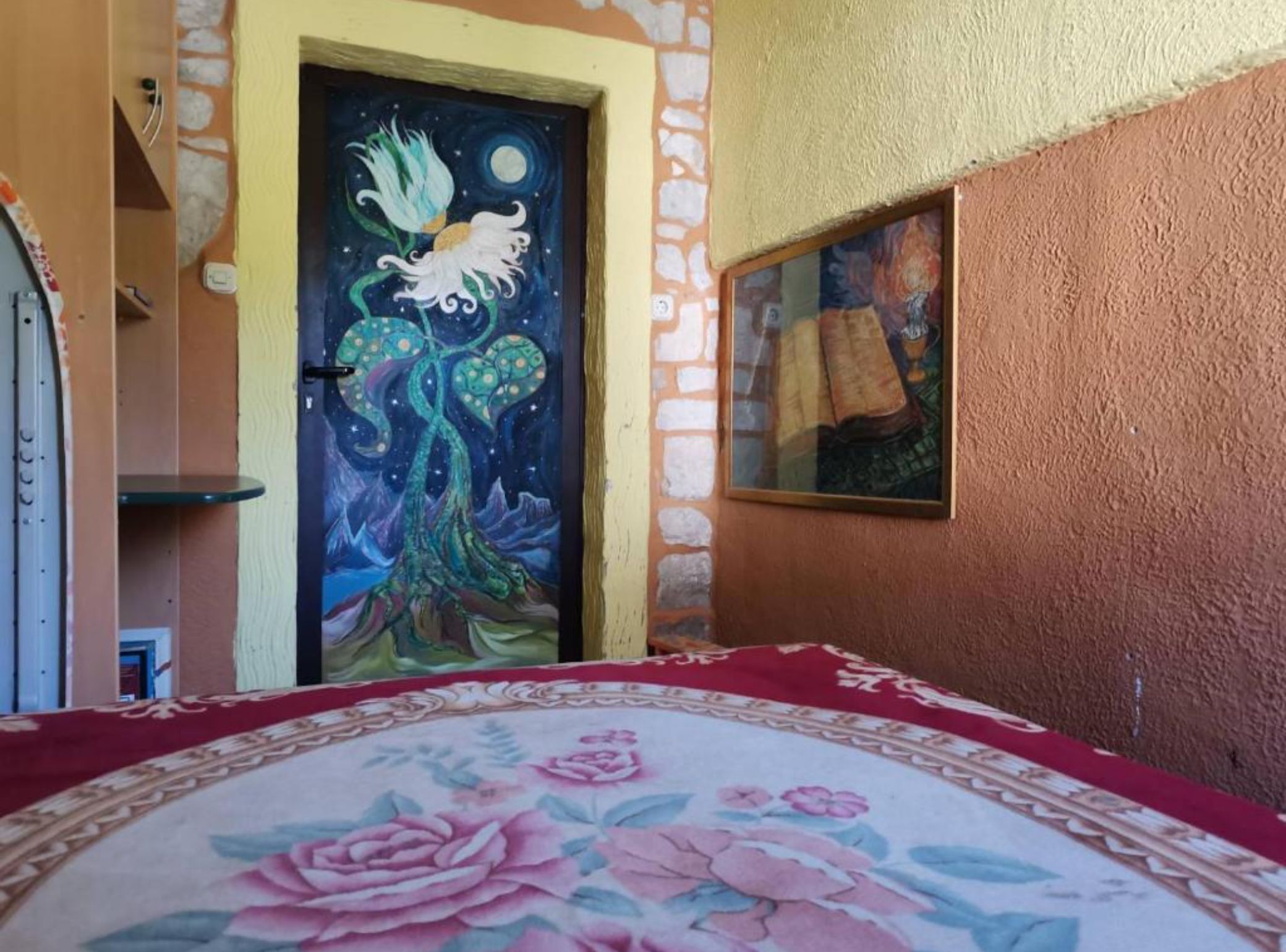 A bedroom with a floral bedspread, a painting of a bird and tree on the door, and a stone wall.