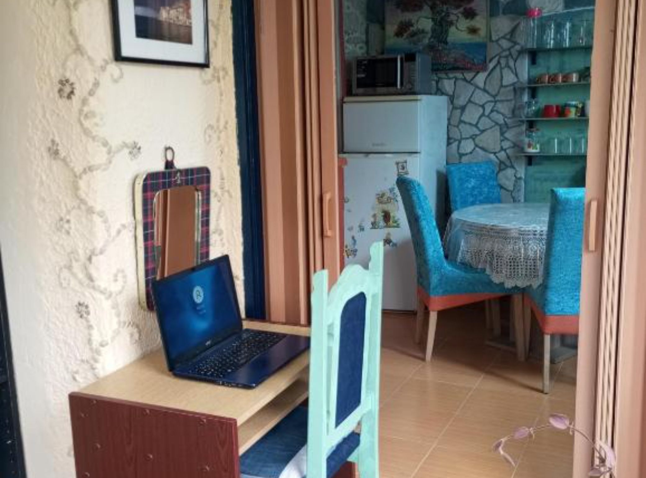 Ein Zimmer mit einem Laptop auf einem Holztisch, einem blauen Stuhl, einem weißen Kühlschrank und einer Steinsäule mit Regalen.