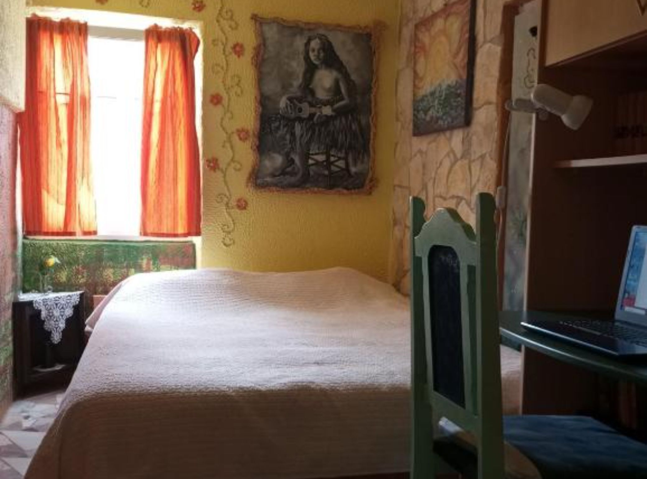 A room with a bed, a desk with a laptop, a window with curtains, and a wall with a painting.