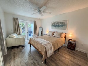 A bedroom with a bed, a seating area, a ceiling fan, and a view through a glass door.