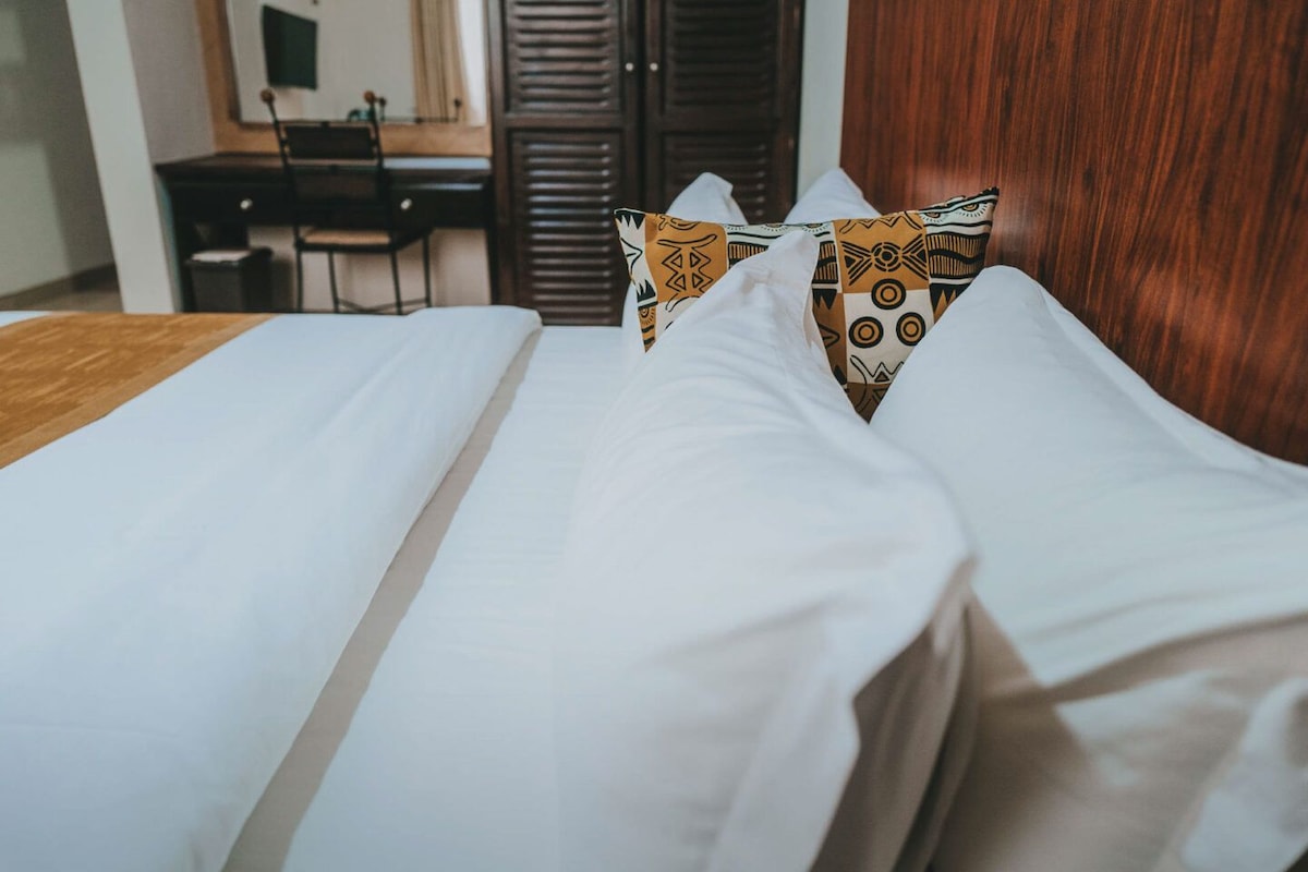 A neatly made bed with white linens and patterned pillows in a room with a wooden headboard and a desk in the background.