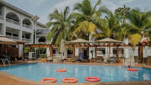 Una piscina con sillones de descanso y sombrillas, rodeada de palmeras y un edificio con balcones.