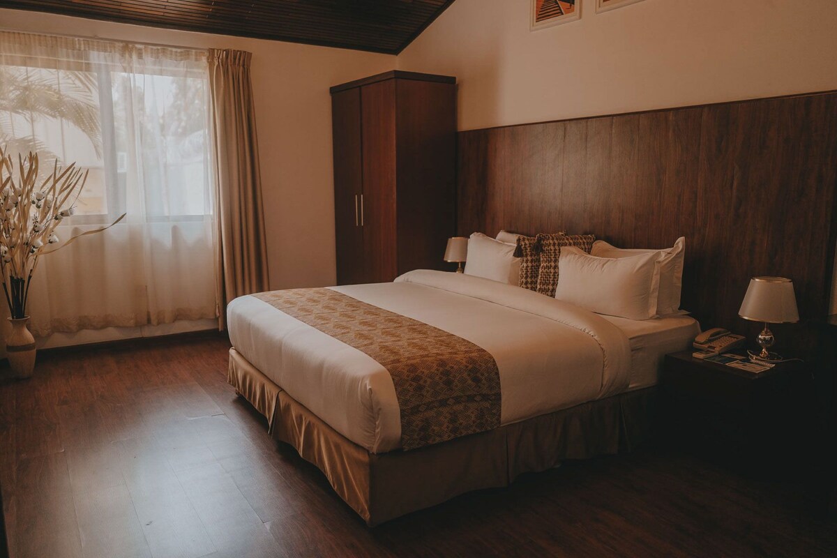 A hotel room with a large bed, a nightstand with a lamp, a wardrobe, and a window with curtains.