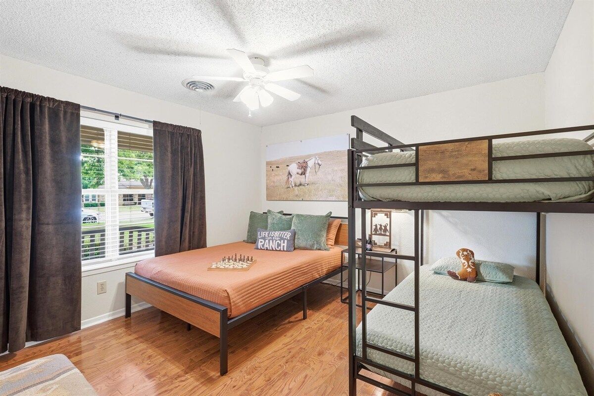 A room with a bunk bed, a single bed, a ceiling fan, a window with curtains, and a painting on the wall.