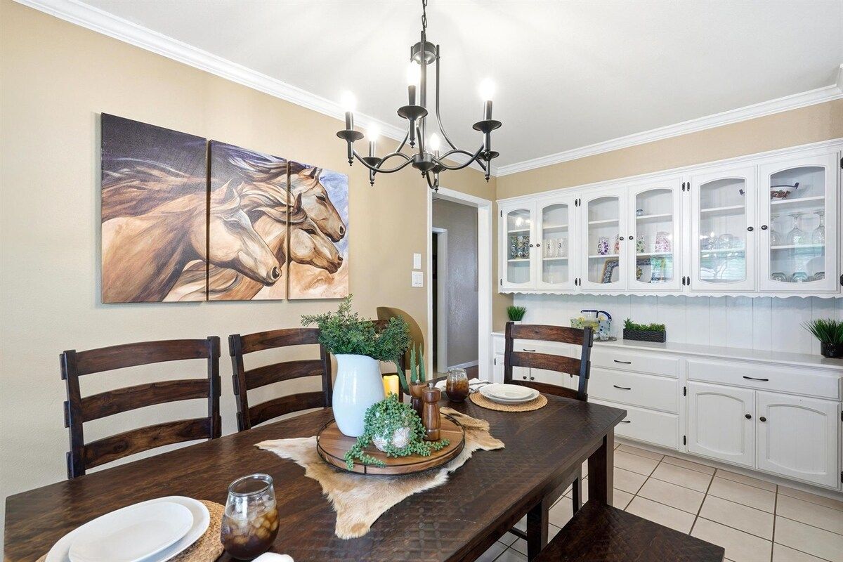 A dining area with a wooden table, chairs, a chandelier, and a painting of horses on the wall.
