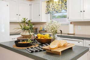 Eine Küche mit weißen Schränken, einer Granit-Arbeitsplatte, einem Holzschneidebrett mit Brot, einer Zitronenschale und einem Blumentopf.