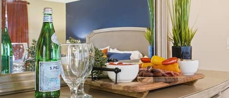 A room with a bed, a wooden side table with a bottle, a glass, and a fruit platter.
