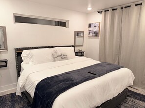 Ein ordentlich bezogenes Bett mit einem Kissen, das 'Willkommen Zuhause' zeigt, ein Nachttisch mit Lampe, ein Fenster mit Blick und ein Vorhang.