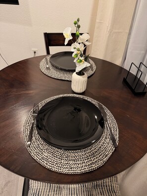 Ein runder Esstisch mit schwarzen Tellern, gewebten Untersetzern, einer weißen Vase mit Blumen und einer schwarzen Servierplatte.