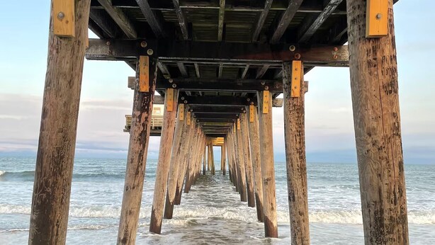 Un muelle de madera que se adentra en el océano, con soportes de madera visibles y una playa de arena al final.