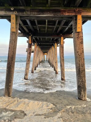 A wooden pier extending into the ocean with visible wooden supports and a sandy beach at the end.